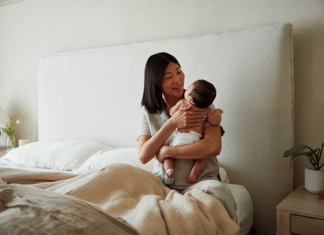 A women sits upright in a bed in a softly lit bedroom holding a baby wearing the Owlet Dream Sock Mint close to her chest.