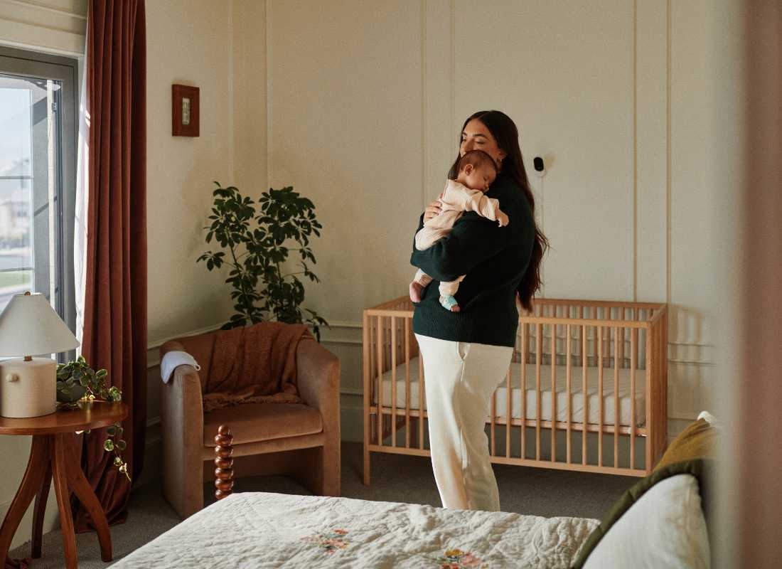 Women hold's a sleeping baby in their arm's whilst standing in the middle of a well lit bedroom